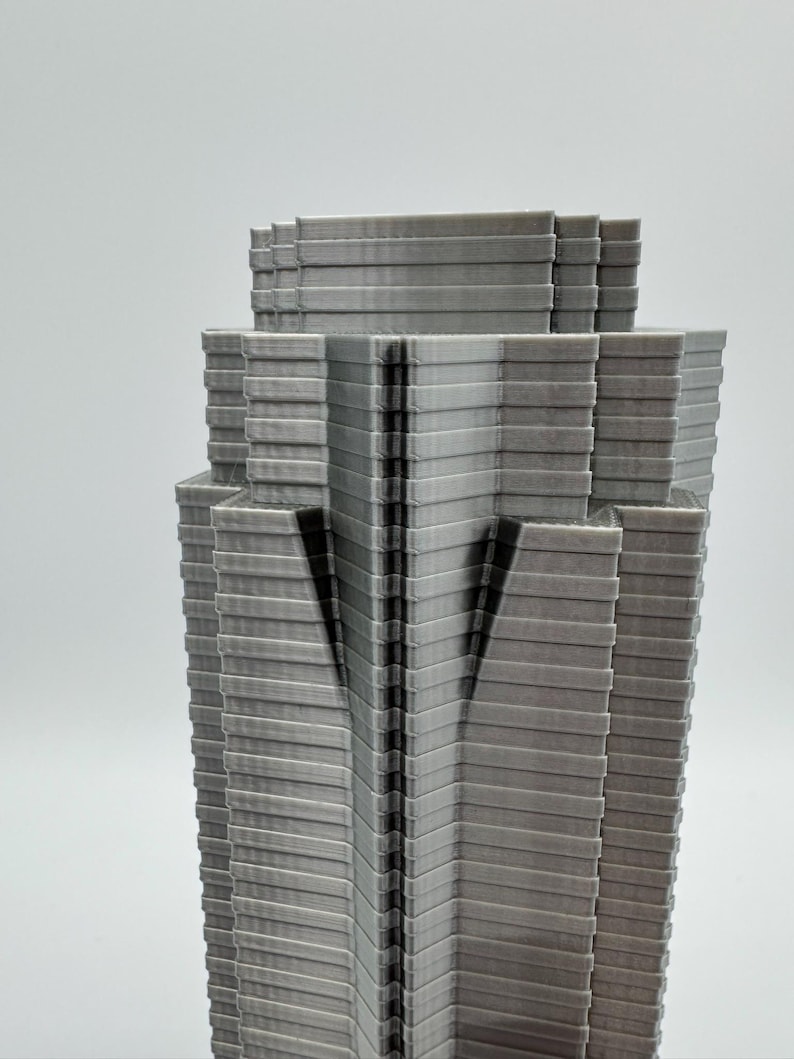 May include: A gray, layered architectural model of a skyscraper. The building has a tiered design with a series of horizontal lines creating a textured appearance. The model is set against a plain white background.