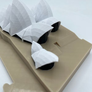 May include: A white 3D printed model of the Sydney Opera House. The model is on a beige base.