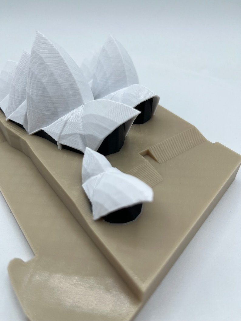 May include: A white 3D printed model of the Sydney Opera House. The model is on a beige base.