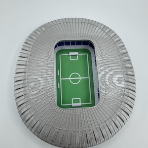 May include: A miniature stadium model with a green field and white markings. The stadium has gray seating and a blue interior. The overall shape is oval, and the model is set against a white background.