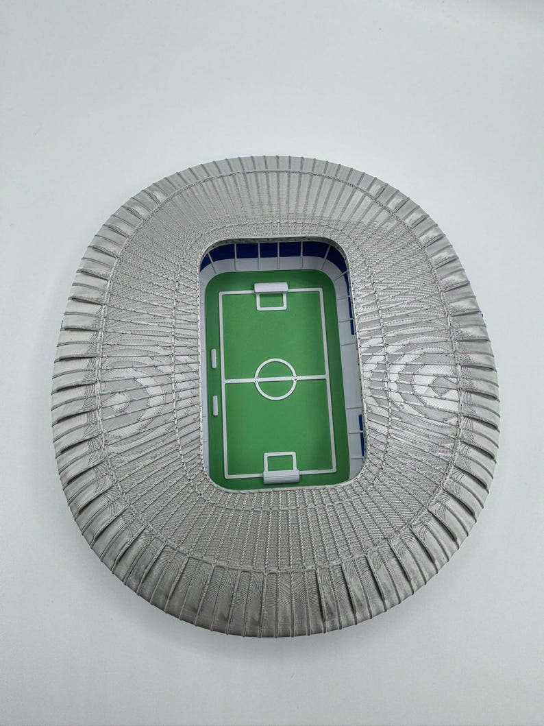 May include: A miniature stadium model with a green field and white markings. The stadium has gray seating and a blue interior. The overall shape is oval, and the model is set against a white background.