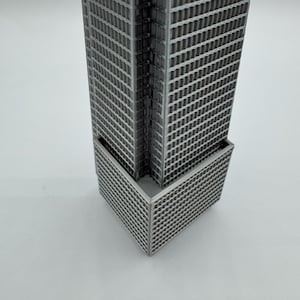 May include: A gray, 3D-printed miniature skyscraper model. The building features a rectangular shape with numerous windows and a detailed facade. The model is a representation of modern architecture.