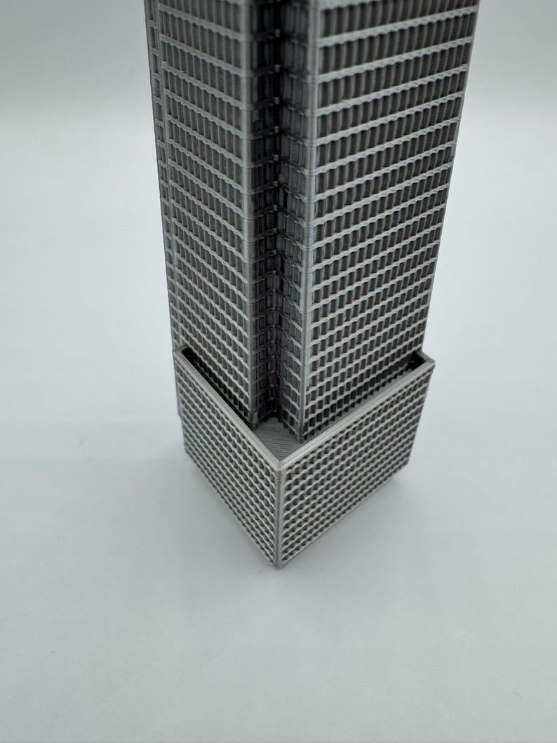 May include: A gray, 3D-printed miniature skyscraper model. The building features a rectangular shape with numerous windows and a detailed facade. The model is a representation of modern architecture.