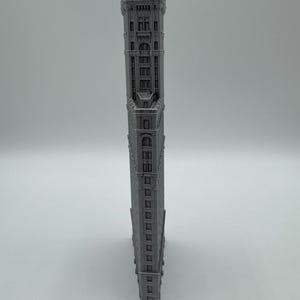 May include: A grey, miniature replica of the Flatiron Building. The model is a detailed representation of the iconic New York City skyscraper, featuring intricate architectural details and a triangular shape. The building is grey.