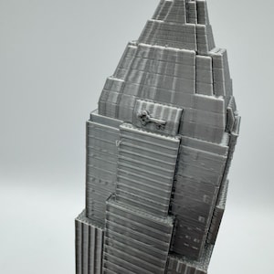 May include: A gray 3D-printed model of a skyscraper, featuring a tiered design with a pointed top. The building has detailed vertical lines and rectangular windows. The model is a solid gray color and stands against a plain background.