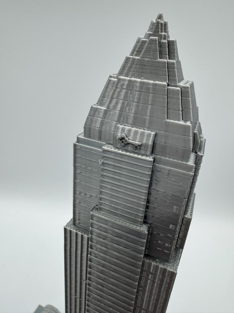 May include: A gray 3D-printed model of a skyscraper, featuring a tiered design with a pointed top. The building has detailed vertical lines and rectangular windows. The model is a solid gray color and stands against a plain background.