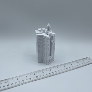 Op de afbeelding: Een wit 3D-geprint model van een hoog gebouw met een decoratieve top. Het model is ongeveer 15 cm hoog en staat op een wit oppervlak.