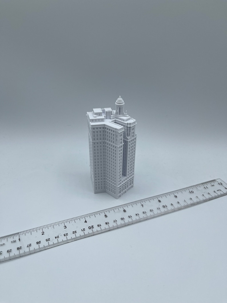 Op de afbeelding: Een wit 3D-geprint model van een hoog gebouw met een decoratieve top. Het model is ongeveer 15 cm hoog en staat op een wit oppervlak.