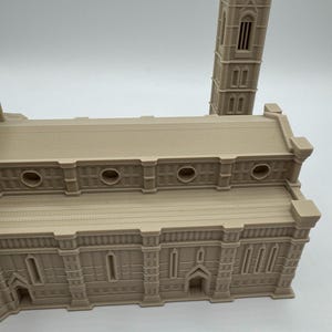 May include: A beige architectural model of a cathedral with a tall bell tower. The model has detailed features including arched windows, circular openings, and a textured roof. The building's facade is intricately designed, showcasing a classic architectural style.
