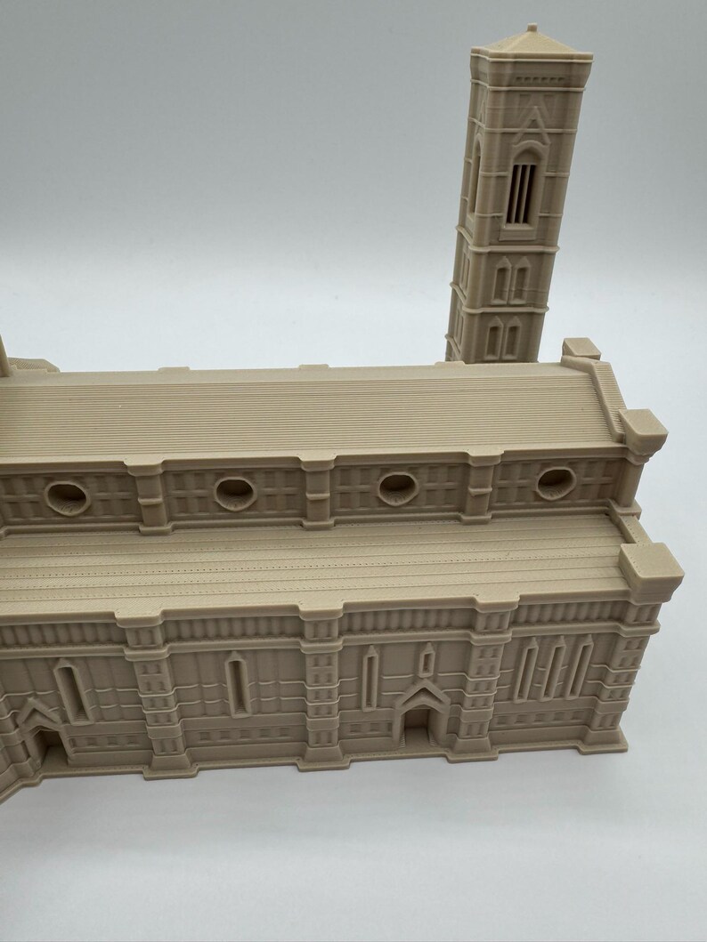 May include: A beige architectural model of a cathedral with a tall bell tower. The model has detailed features including arched windows, circular openings, and a textured roof. The building's facade is intricately designed, showcasing a classic architectural style.