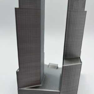 Op de afbeelding: Een 3D-geprint model van twee hoge gebouwen, waarvan er &eacute;&eacute;n iets hoger is dan de andere, met een plat dak dat ze verbindt. De gebouwen zijn gemaakt van grijs plastic en hebben een getextureerd oppervlak.