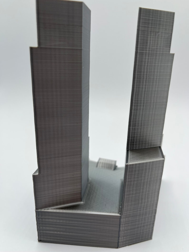 Op de afbeelding: Een 3D-geprint model van twee hoge gebouwen, waarvan er &eacute;&eacute;n iets hoger is dan de andere, met een plat dak dat ze verbindt. De gebouwen zijn gemaakt van grijs plastic en hebben een getextureerd oppervlak.