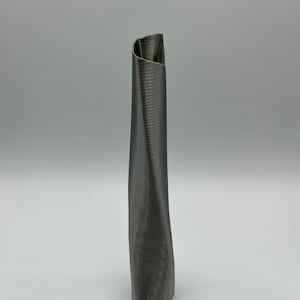 Op de afbeelding: Een grijze, 3D-geprinte vaas met een uniek, gedraaid ontwerp. De vaas is hoog en slank, met een smalle opening aan de bovenkant.