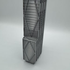 May include: A silver-colored 3D printed model of a skyscraper featuring a distinct X-braced structural exterior design. The tall, rectangular building replica displays vertical lines and a repeating diamond-shaped truss pattern along its facade.