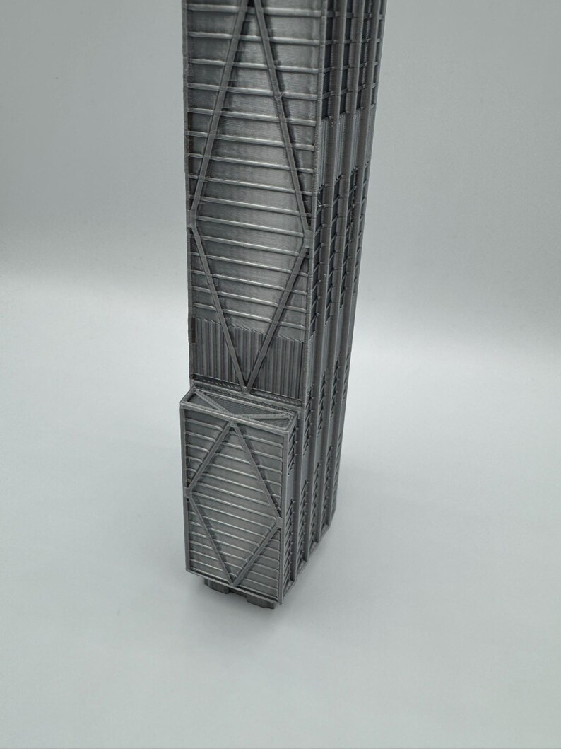 May include: A silver-colored 3D printed model of a skyscraper featuring a distinct X-braced structural exterior design. The tall, rectangular building replica displays vertical lines and a repeating diamond-shaped truss pattern along its facade.