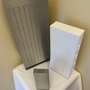 May include: Three miniature skyscraper models made of gray and white plastic LEGO bricks. The models are arranged on a white tablecloth. The largest model is gray and the two smaller models are white and gray.