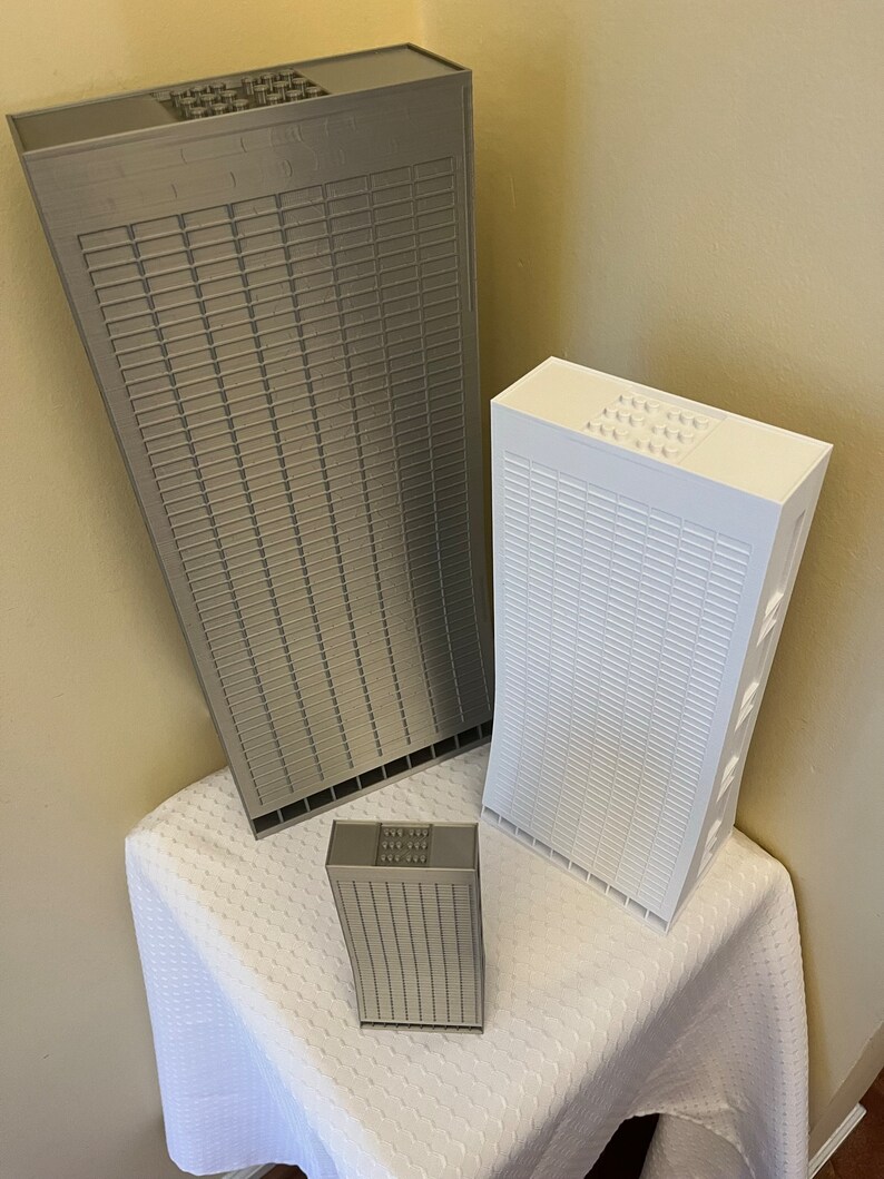 May include: Three miniature skyscraper models made of gray and white plastic LEGO bricks. The models are arranged on a white tablecloth. The largest model is gray and the two smaller models are white and gray.