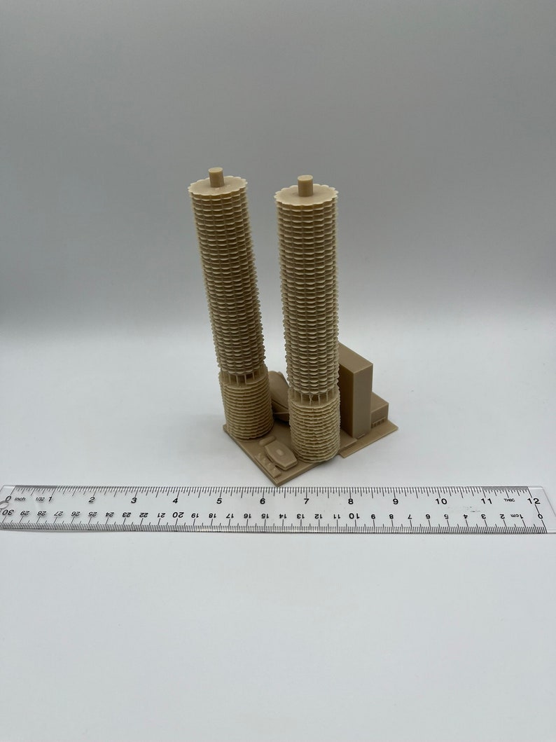 May include: A 3D printed model of a pair of tan-colored buildings with a unique architectural design. The model is approximately 7 inches tall and is sitting on a white surface.