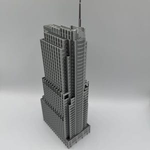 May include: A gray, detailed model of a skyscraper with a tall antenna. The building has many windows and a stepped design, typical of Art Deco architecture. The model is set against a plain background.