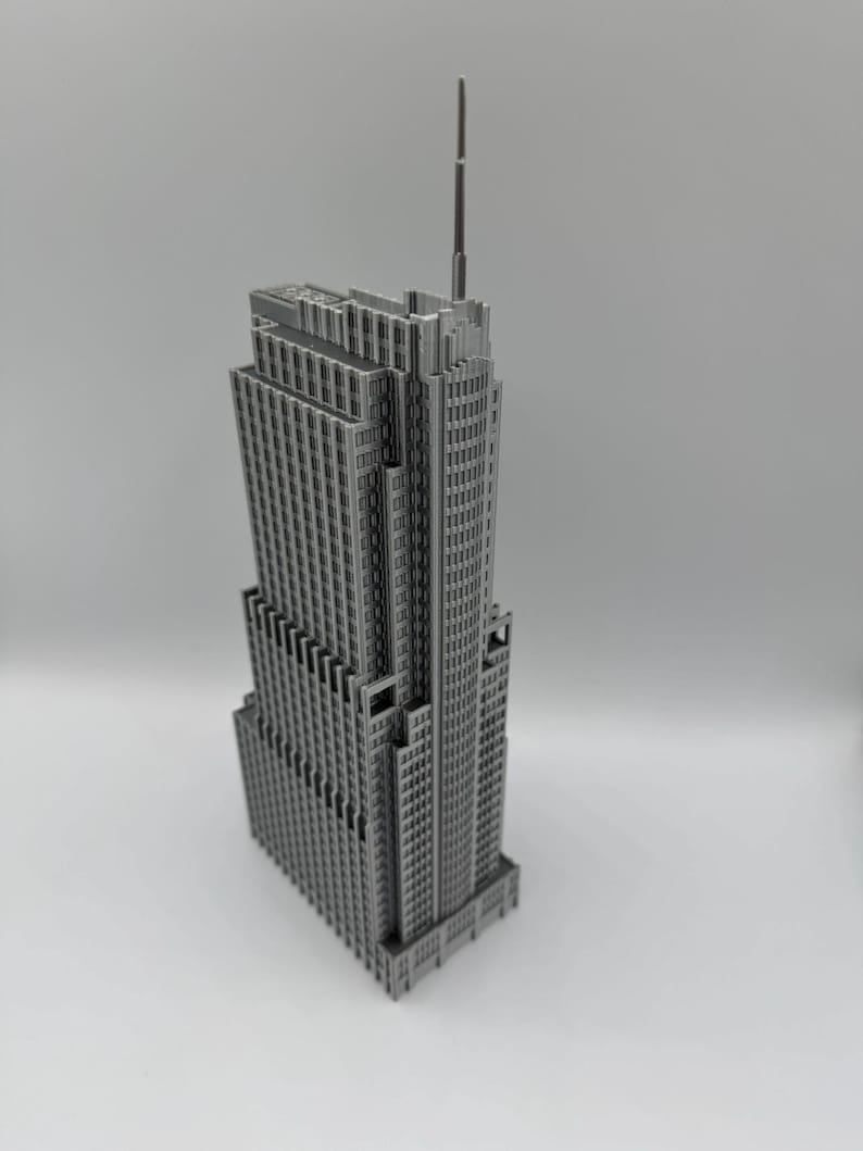 May include: A gray, detailed model of a skyscraper with a tall antenna. The building has many windows and a stepped design, typical of Art Deco architecture. The model is set against a plain background.