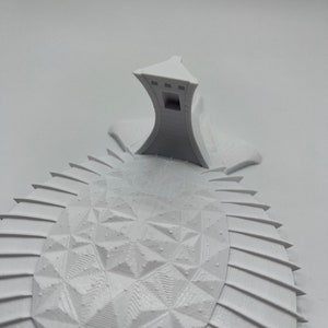 May include: A white 3D printed model of a building with a geometric design. The building has a tall, pointed roof and a series of windows on the sides. The model is sitting on a white surface.