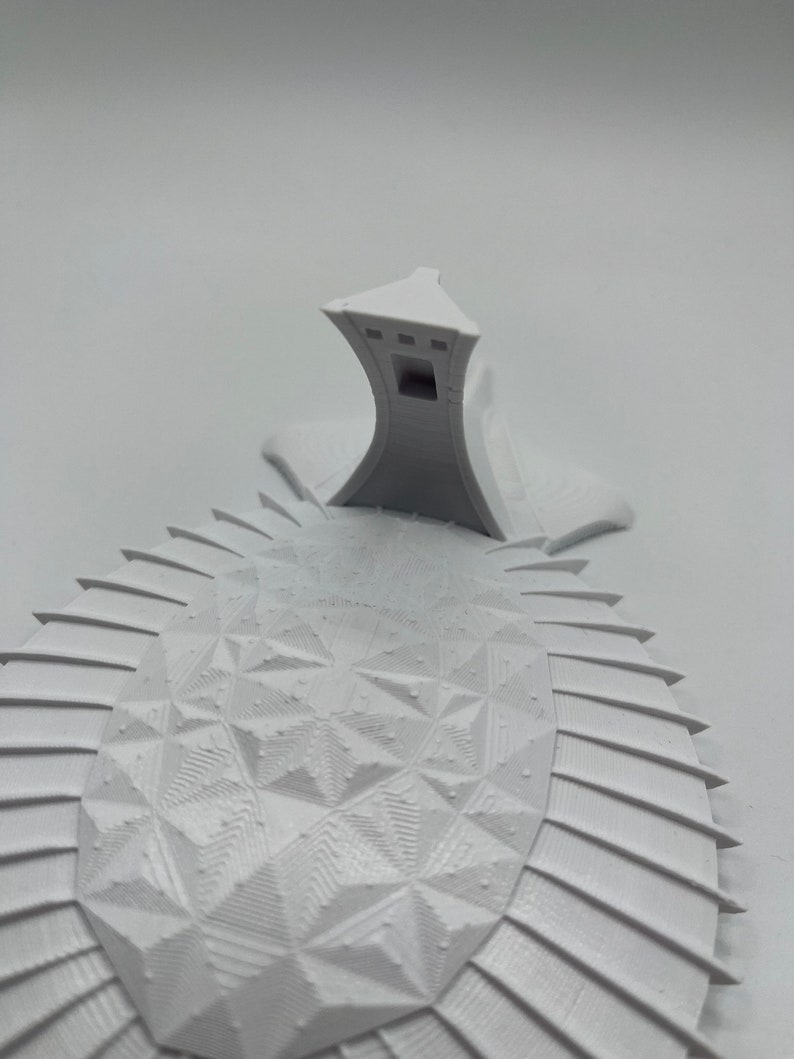 May include: A white 3D printed model of a building with a geometric design. The building has a tall, pointed roof and a series of windows on the sides. The model is sitting on a white surface.