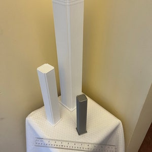 Op de afbeelding: Drie witte 3D-geprinte modellen van wolkenkrabbers, &eacute;&eacute;n groot en twee kleinere, op een wit tafelkleed met een liniaal als schaal.