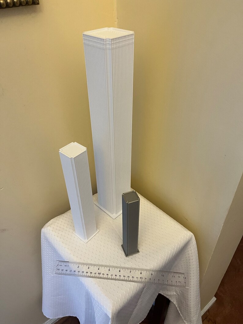 Op de afbeelding: Drie witte 3D-geprinte modellen van wolkenkrabbers, &eacute;&eacute;n groot en twee kleinere, op een wit tafelkleed met een liniaal als schaal.