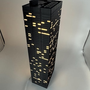 Peut inclure: Une lampe noire en forme de gratte-ciel avec des fen&ecirc;tres &eacute;clair&eacute;es. La lampe est rectangulaire et poss&egrave;de un dessus plat avec une petite structure. Les fen&ecirc;tres sont de tailles et de formes vari&eacute;es, &eacute;mettant une lumi&egrave;re jaune chaude. Un c&acirc;ble blanc sort de la base.