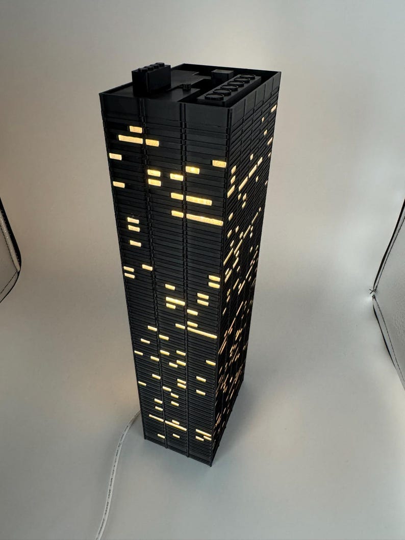 Peut inclure: Une lampe noire en forme de gratte-ciel avec des fen&ecirc;tres &eacute;clair&eacute;es. La lampe est rectangulaire et poss&egrave;de un dessus plat avec une petite structure. Les fen&ecirc;tres sont de tailles et de formes vari&eacute;es, &eacute;mettant une lumi&egrave;re jaune chaude. Un c&acirc;ble blanc sort de la base.