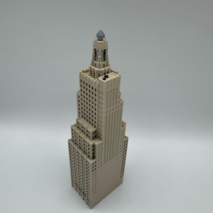 May include: A beige miniature skyscraper model with a detailed facade, featuring numerous windows and a pointed top with a dark gray gem. The building has a tiered design and is set against a plain background.