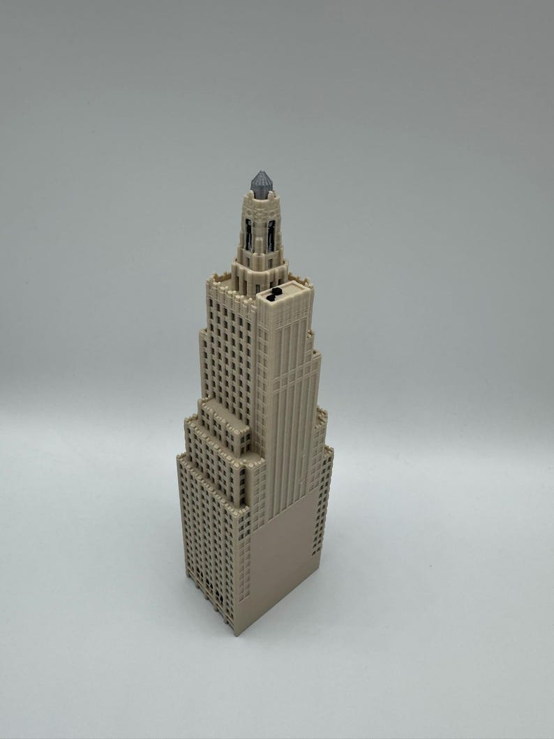 May include: A beige miniature skyscraper model with a detailed facade, featuring numerous windows and a pointed top with a dark gray gem. The building has a tiered design and is set against a plain background.