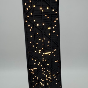 May include: A black architectural model of a skyscraper with illuminated windows. The model features a detailed structural design and is lit with warm, yellow lights, creating a nighttime cityscape effect. The building is angled slightly, showcasing its height and intricate details.