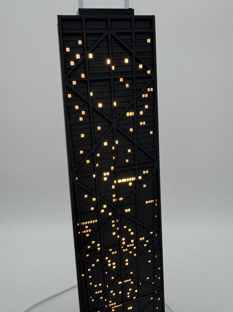 May include: A black architectural model of a skyscraper with illuminated windows. The model features a detailed structural design and is lit with warm, yellow lights, creating a nighttime cityscape effect. The building is angled slightly, showcasing its height and intricate details.
