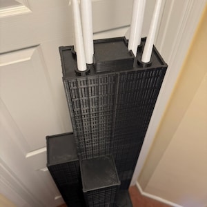 May include: A black and white 3D-printed model of a skyscraper, with detailed window patterns and white cylindrical structures on the roof. The model is a replica of a famous building.