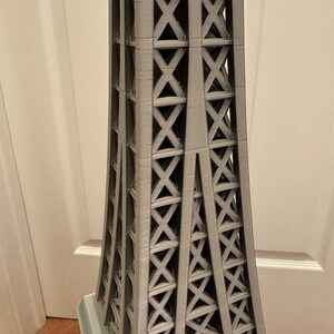May include: A gray Eiffel Tower model with a detailed lattice design. The tower's structure is visible, showcasing the intricate crisscross pattern. The model is a miniature representation of the iconic landmark.