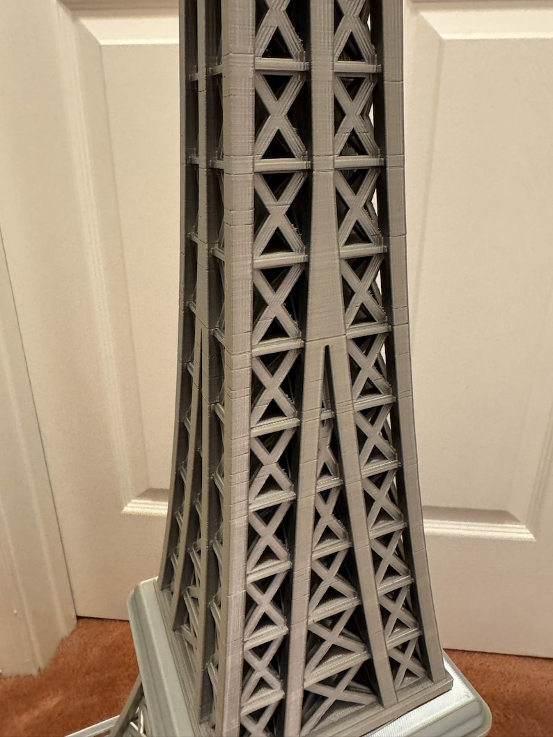 May include: A gray Eiffel Tower model with a detailed lattice design. The tower's structure is visible, showcasing the intricate crisscross pattern. The model is a miniature representation of the iconic landmark.