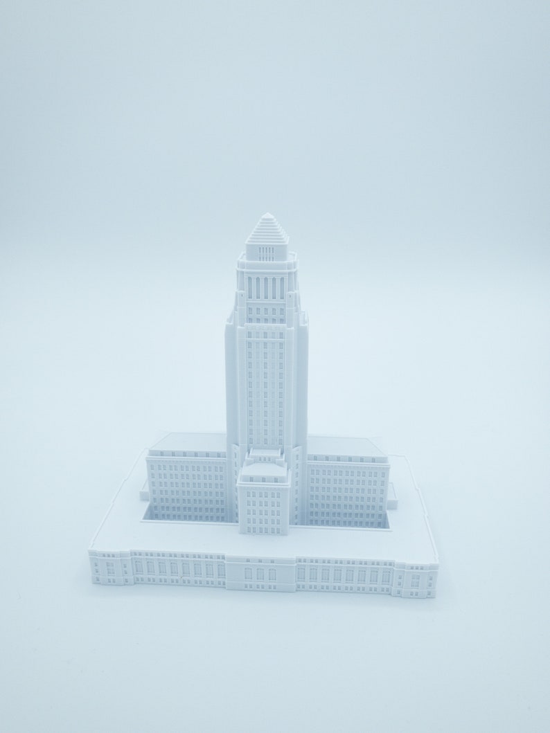 May include: A white 3D printed model of a city hall building. The building has a tall tower with a pointed roof and a rectangular base with many windows.