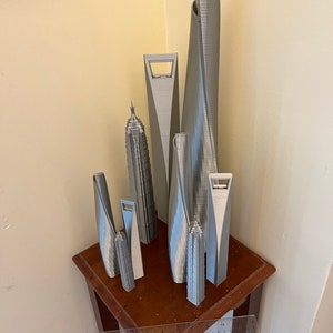 May include: A collection of seven silver 3D printed miniature skyscrapers on a wooden table. The skyscrapers are of varying heights and designs, with some featuring unique architectural details.