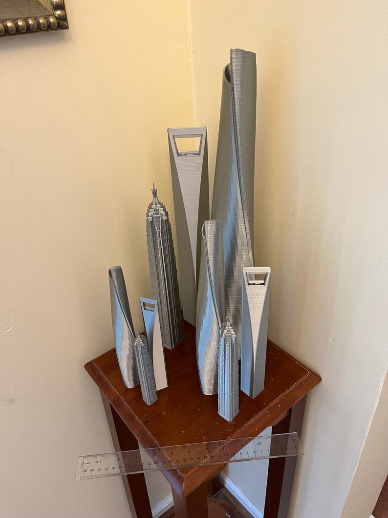 May include: A collection of seven silver 3D printed miniature skyscrapers on a wooden table. The skyscrapers are of varying heights and designs, with some featuring unique architectural details.