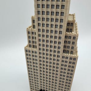 May include: A beige 3D-printed model of a skyscraper with numerous windows and a tiered design. The building has a detailed facade with a textured appearance, and a dark base. The model is set against a plain white background.