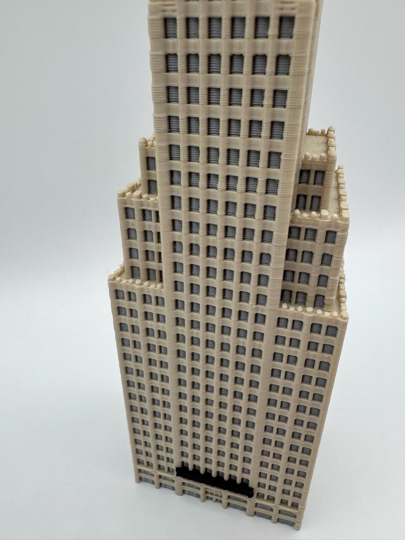May include: A beige 3D-printed model of a skyscraper with numerous windows and a tiered design. The building has a detailed facade with a textured appearance, and a dark base. The model is set against a plain white background.