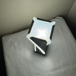 May include: A white and black geometric lamp with a square base and a rectangular light source. The lamp is turned on and illuminating the surrounding area.