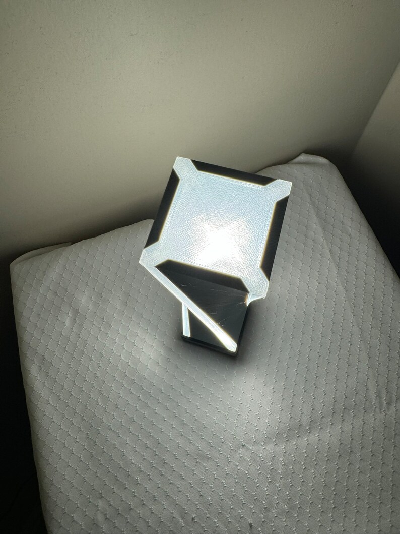 May include: A white and black geometric lamp with a square base and a rectangular light source. The lamp is turned on and illuminating the surrounding area.