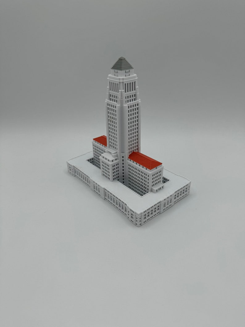 May include: A white miniature model of the Los Angeles City Hall building with a red roof. The building has a tall tower with the words "City Hall" on the side.
