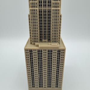 May include: A beige miniature of a skyscraper with a rectangular base and a tiered top. The building has numerous small, dark windows and vertical lines, creating a detailed architectural model. The overall design is a simplified representation of a famous building.