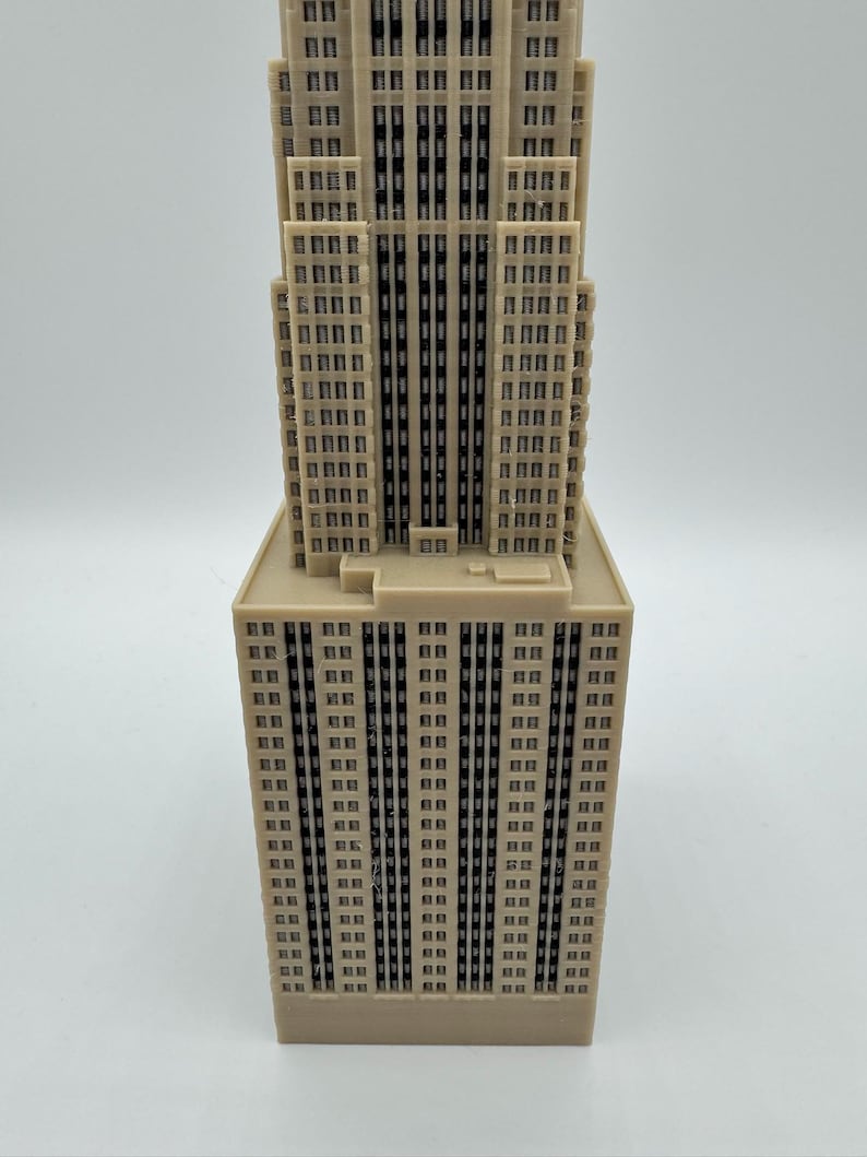 May include: A beige miniature of a skyscraper with a rectangular base and a tiered top. The building has numerous small, dark windows and vertical lines, creating a detailed architectural model. The overall design is a simplified representation of a famous building.