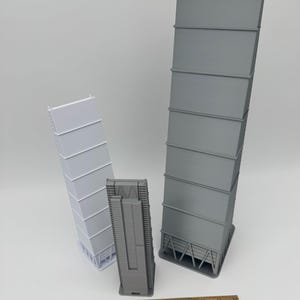 May include: Three 3D-printed industrial models in varying sizes. The largest is gray, the medium is also gray, and the smallest is white. A wooden ruler is at the bottom, providing scale. The models have a rectangular shape with horizontal lines.