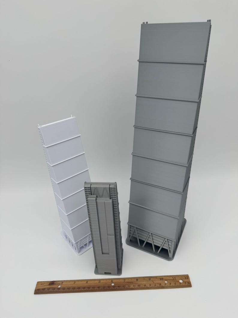 May include: Three 3D-printed industrial models in varying sizes. The largest is gray, the medium is also gray, and the smallest is white. A wooden ruler is at the bottom, providing scale. The models have a rectangular shape with horizontal lines.