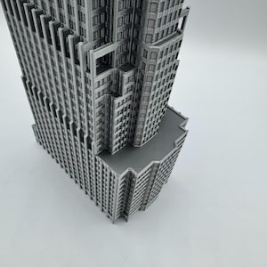 May include: A detailed, gray miniature skyscraper model. The building features multiple levels, windows, and architectural details. The model is set against a plain white background, highlighting its intricate design.